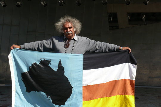 Maurie Japarta Ryan sur la place de la Fédération à Melbourne, lors de son deuxième «meeting solitaire». Dans ses bras, le drapeau de son parti First Nations, avec à droite les quatre couleurs utilisées dans les peintures corporelles lors des cérémonies traditionnelles. (Radio Australia/Caroline Lafargue)